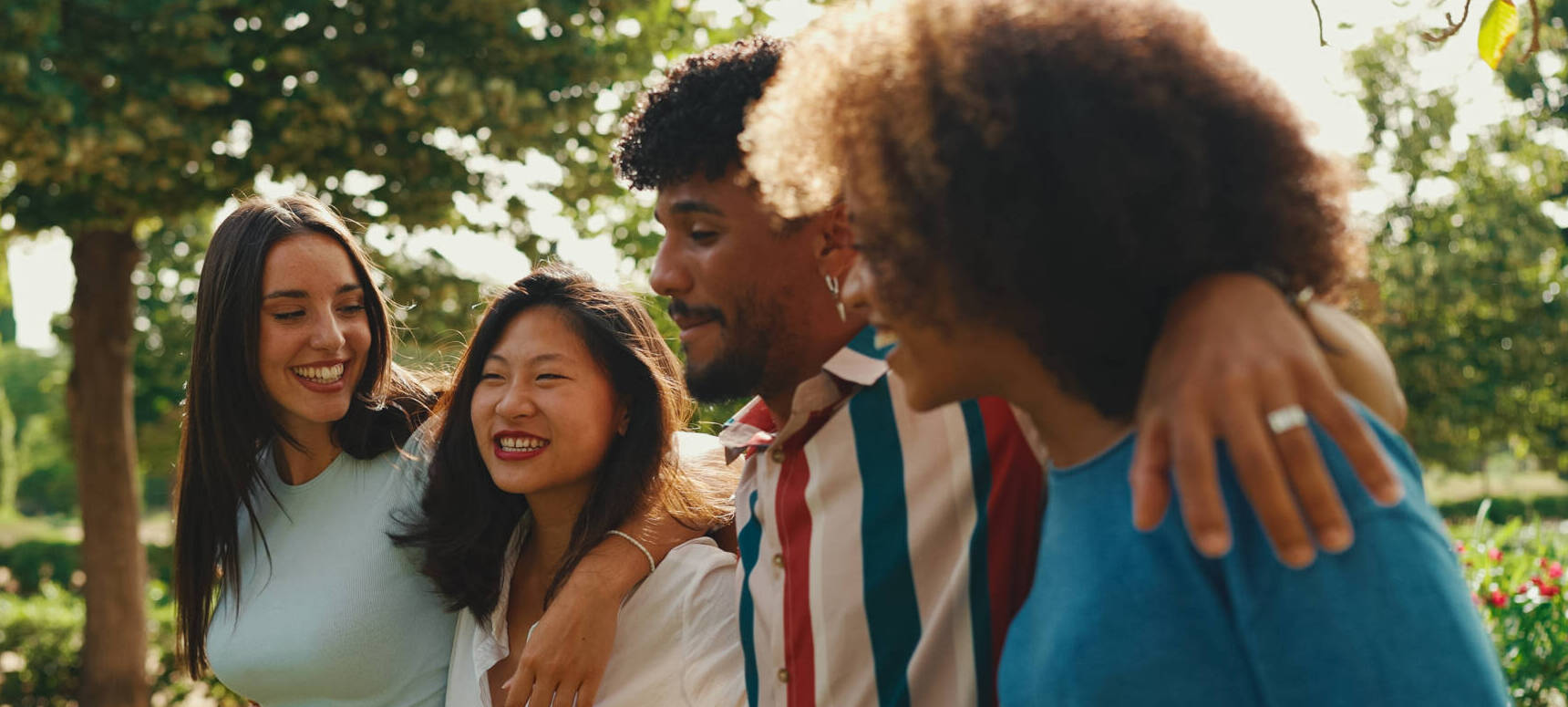 Happy Multiethnic Young People Walk Embracing On Summer Day Outdoors. Group Of Friends Are Talking And Laughing Merrily While Walking Along Path In City Park